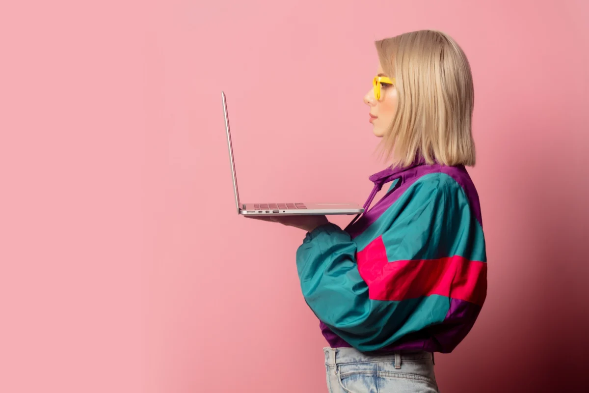 Młoda kobieta w żółtych okularach na różowym tle ubrana w stylu lat 90 trzymająca przed sobą laptopa i patrząca na jego ekran